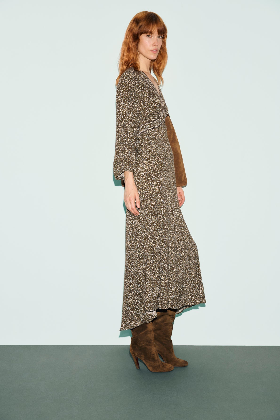 A woman with red hair wearing a robe longue in tissu fluide with an imprimé abstrait and brown suede boots stands against a light blue background, looking at the camera.