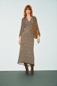 A woman with red hair wears a long, patterned brown dress in tissu fluide, brown knee-high boots, and carries a brown suede shoulder bag, standing against a plain light background.