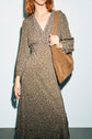A person with shoulder-length hair wears a robe longue à imprimé abstrait en tissu fluide and carries a tan suede shoulder bag, standing against a plain light background. Their face is partially out of the frame.