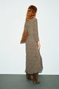 A person with wavy, shoulder-length red hair stands facing away, wearing a robe longue à imprimé abstrait in tissu fluide, complemented by brown high-heeled boots and a brown shoulder bag against a light background.