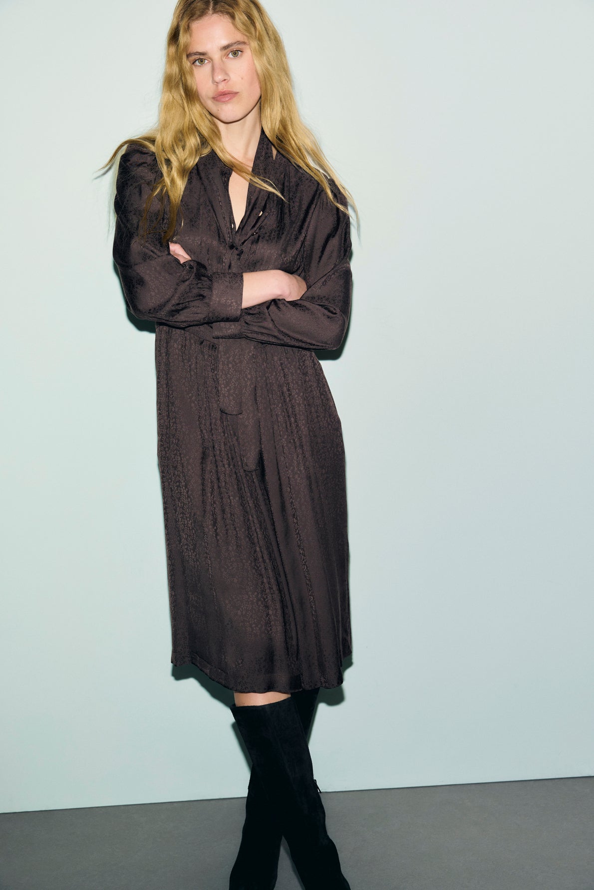 A person with long blond hair stands against a plain light background, wearing a knee-length, long-sleeve robe jacquard dress and black boots, with arms crossed and a neutral expression.