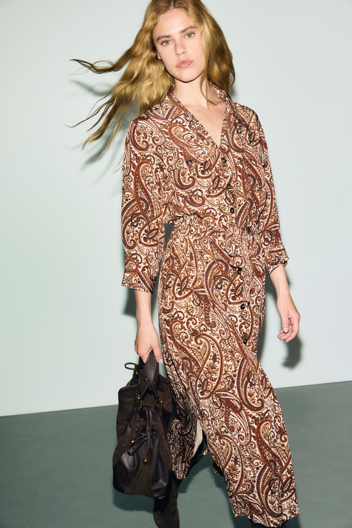 A woman with long, wavy blonde hair exudes allure bohème as she wears a brown robe chemise adorned with a delicate imprimé cachemire and holds a dark handbag, walking against a light background.