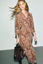 A woman with long, wavy blonde hair exudes allure bohème as she wears a brown robe chemise adorned with a delicate imprimé cachemire and holds a dark handbag, walking against a light background.