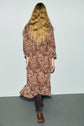 A person with long, wavy blonde hair is shown from behind, wearing a brown robe chemise à imprimé cachemire and knee-high brown boots, standing on a gray floor against a light background for an effortlessly chic allure bohème.
