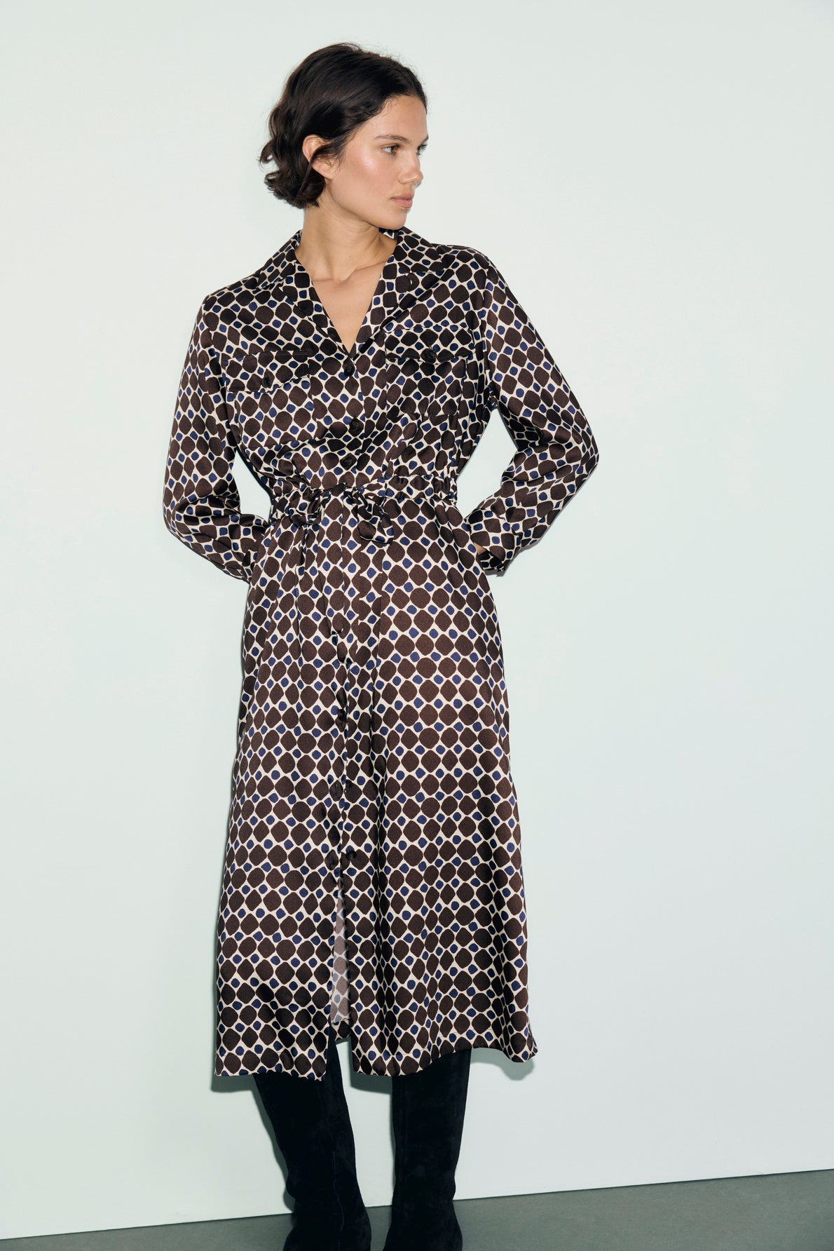 A person wearing a long, patterned brown and white robe midi with a collar, long sleeves, and a ceinture à nouer at the waist stands against a plain light background, looking to the side.