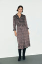 A woman stands against a plain wall, wearing a brown, long-sleeve, button-down robe midi with a white imprimé graphique and black knee-high boots.