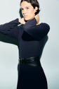 Person with short dark hair wearing a long-sleeved, fitted robe midi en maille côtelée bleu marine and a wide black belt, adjusting their collar and looking slightly to the side against a plain light background.