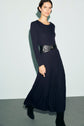 A woman wearing a long-sleeved, bleu marine robe midi in maille côtelée with a wide black belt and black boots stands against a plain light background.