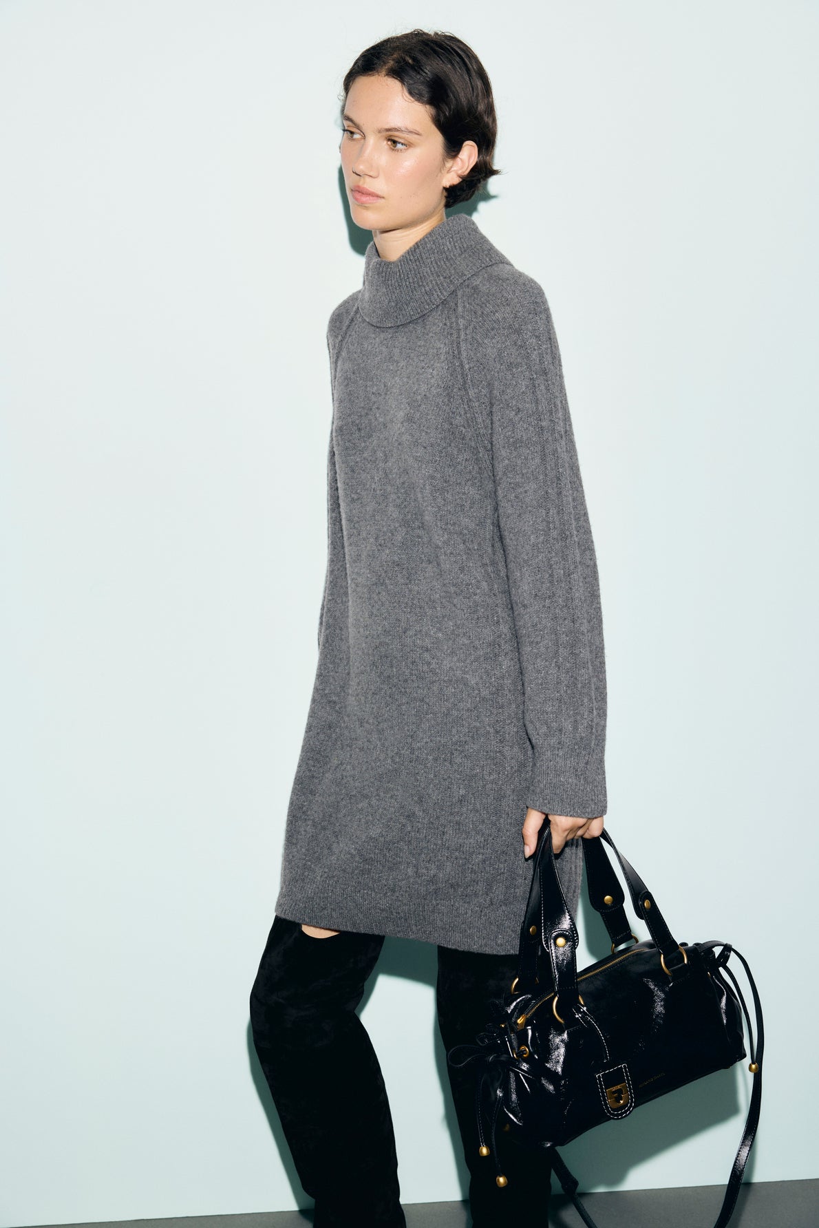 A person wearing a long gray robe en laine and black boots stands against a light background, holding a shiny black handbag.