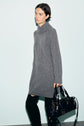 A person wearing a long gray robe en laine and black boots stands against a light background, holding a shiny black handbag.