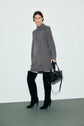 Person wearing a robe en laine gris à manches longues avec un col roulé ouvert, une robe courte associée à des bottes noires à talons au-dessus du genou, tenant un sac à main noir et posant devant un fond clair et uni.