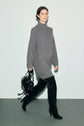 A person wearing a long gray turtleneck sweater dress with a hint of col roulé ouvert detail and black over-the-knee boots walks while holding a black handbag against a plain light-colored background.