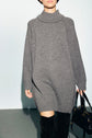A person wearing a long, loose gray robe en laine turtleneck sweater dress, black knee-high boots, and holding a black handbag, photographed against a plain background.
