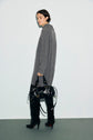 A person wearing a long gray sweater with a col roulé ouvert, black leggings, and black high-heeled boots stands against a plain background, holding a shiny black handbag.
