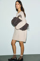 A woman wearing a light beige robe en laine à manches longues, brown socks, and black loafers holds a large brown knit bag under her arm, standing against a pale blue background.