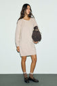 A woman in a beige robe en laine avec manches longues stands holding a brown handbag, looking slightly to the side against a plain light background. She pairs her robe courte with brown loafers and beige socks for an effortlessly chic look.