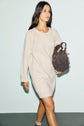 A woman in a beige knitted robe en laine walks while carrying a brown handbag. She has long dark hair and wears neutral-colored socks. The robe courte features manches longues, adding to the cozy, elegant look against the plain, light-colored background.