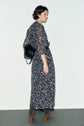 A person with short dark hair wears a robe longue à motif à fleurs, brown suede boots, and carries a black handbag over one shoulder, posing against a plain light background.