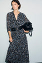 Woman in a dark robe longue with a motif à fleurs and a black belt, holding a large black leather bag under her arm, stands against a plain light background.