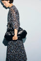 Woman in a robe longue à motif à fleurs holds a large black leather bag under her arm, standing against a pale background. She is facing left, with short hair, and is partially in shadow.