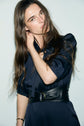 Woman with long brown hair wearing a robe élégante femme in a deep, dark shade and a wide black belt, looking at the camera with her head tilted and one hand touching her hair, standing against a plain light background.