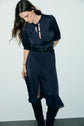 Woman with long brown hair looks down, wearing a navy silk dress with a bow at the neck, short sleeves, a black belt, and knee-high black boots—an élégante robe midi satin in encre profond—standing against a plain white background.