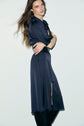 A person with long brown hair stands sideways, wearing a navy blue, long-sleeved, belted robe élégante femme with a front slit and black boots against a plain light background.