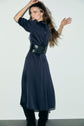 Woman with long brown hair poses in a navy belted robe midi satin and black boots, standing against a plain white wall. She looks over her shoulder with one arm raised and the other bent, exuding the timeless allure of une robe élégante femme.