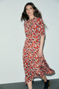 A woman with long brown hair walks, wearing a robe imprimé floral rouge de style midi avec un col à nouer et des bottines noires, against a plain white background.