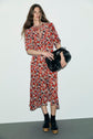 Woman standing against a plain wall, wearing a robe midi imprimé floral rouge with black platform sandals and black socks, holding a large black handbag, with long wavy hair and a relaxed smile.