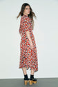 A woman with long brown hair stands sideways, wearing a robe midi imprimé floral rouge and black platform sandals with wooden heels, against a plain light background.