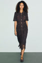 A person with curly hair wears a dark, short-sleeve, button-down robe dentelle midi dress with patch pockets and matching shoes, standing against a plain light background.