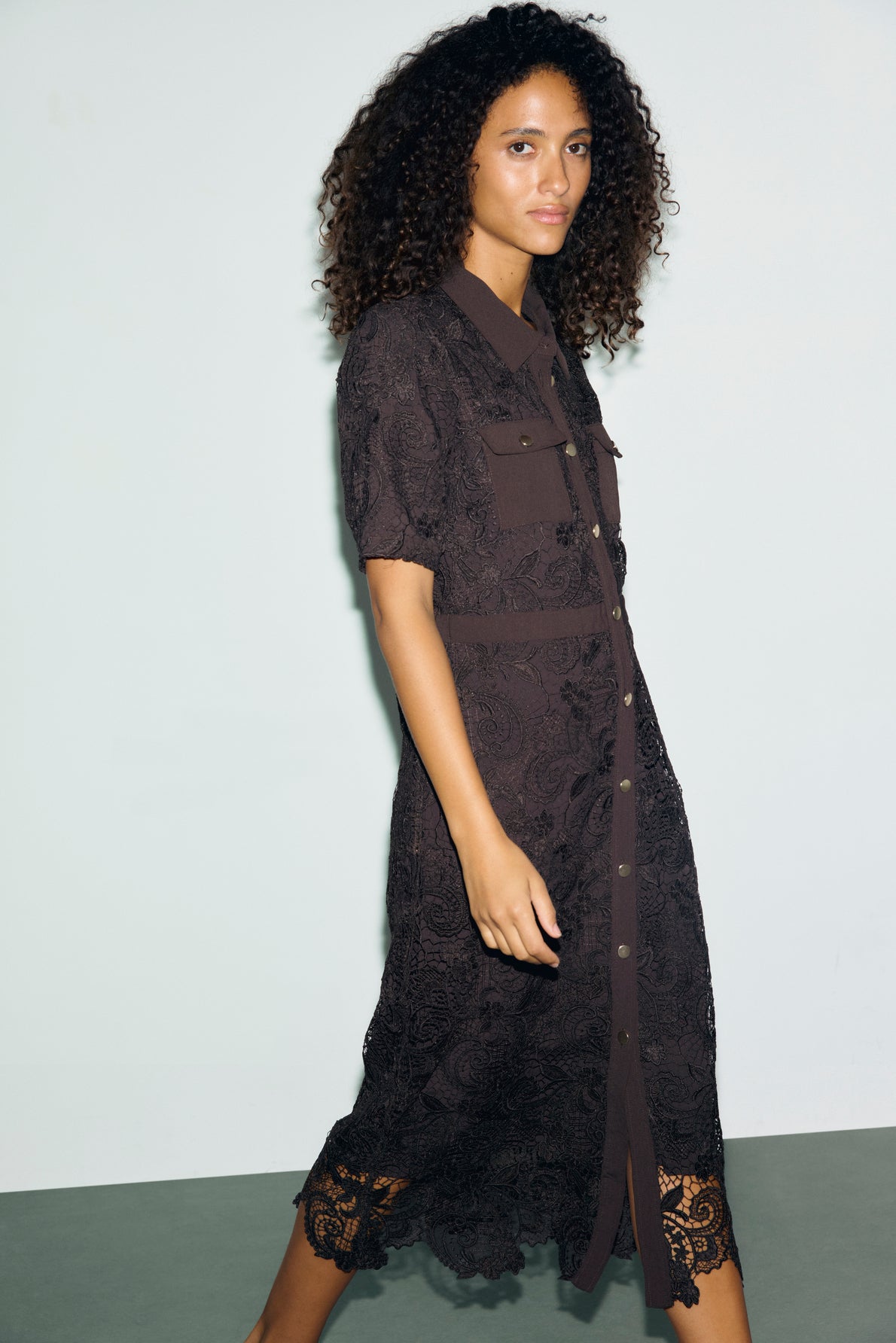 A woman with curly hair wears a robe midi cintrée en dentelle, featuring short sleeves, chest pockets, and a button-down front, as she stands against a plain light background.