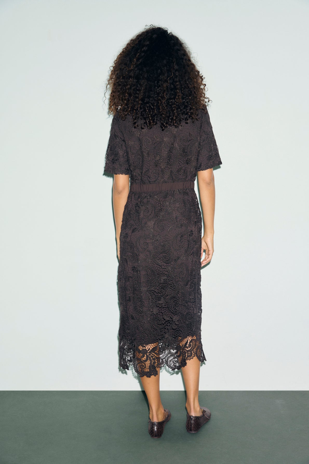 A person with curly hair stands facing away, wearing a robe cintrée en dentelle noire à manches courtes et longueur genou, assortie de chaussures marron, sur un fond uni et clair.