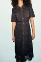 A woman with curly hair wears a black lace robe midi, short-sleeve, button-down dress with front pockets and metal snap buttons, standing against a plain light background. Her face is not visible in the image.