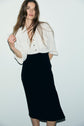 Woman with long brown hair wears a loose white button-up shirt tucked into a high-waisted black robe midi élégante, standing against a plain white background.
