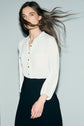 A person in a white blouse and black skirt stands against a plain light background, with their long brown hair blowing to the side and a neutral expression on their face, evoking the timeless appeal of a robe bicolore.