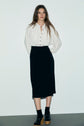 A person stands against a plain white wall, wearing a robe midi bicolore with a sophisticated touch—its white top seamlessly tucked into the black skirt. They complete the look with black socks and black platform sandals, epitomizing an effortlessly chic style.