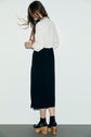 A woman stands against a plain white wall, facing away. She wears a long-sleeved white top paired with a robe bicolore—a black skirt that creates a striking contrast—along with black socks and wooden platform sandals. Her long hair flows down her back.