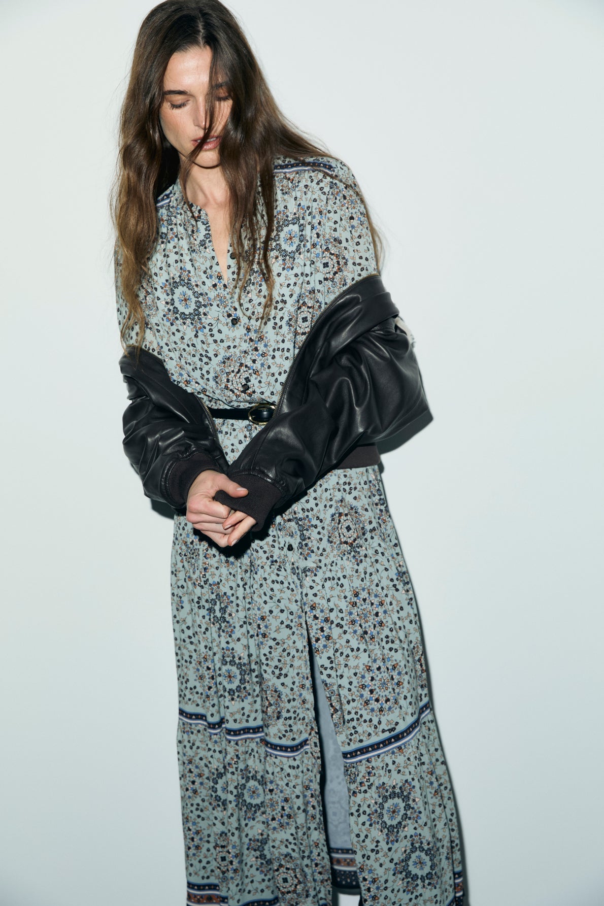 A woman in a long, imprimé floral robe boutonnée with a black jacket draped over her shoulders stands against a plain light background, looking down with her hands clasped.