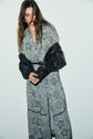 A woman in a long, imprimé floral robe boutonnée with a black jacket draped over her shoulders stands against a plain light background, looking down with her hands clasped.
