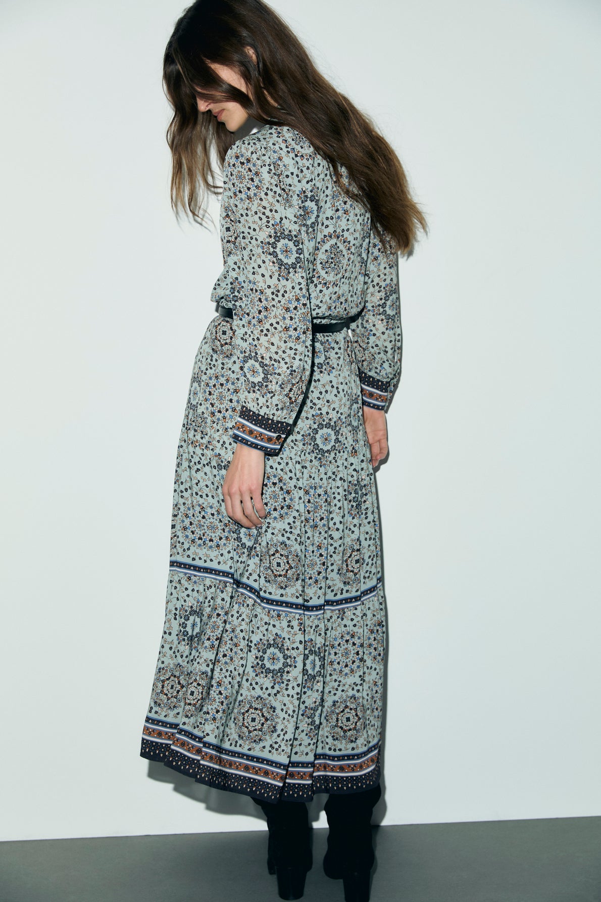 Woman with long brown hair stands against a white wall, looking down. She wears a robe longue imprimé floral in blue-gray tones and black boots. Her face is partially obscured by her hair.