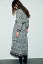 Woman with long brown hair stands against a white wall, looking down. She wears a robe longue imprimé floral in blue-gray tones and black boots. Her face is partially obscured by her hair.