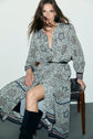A woman with long hair, wearing a robe longue imprimé floral and black boots, sits sideways on a chair against a plain background, looking directly at the camera.