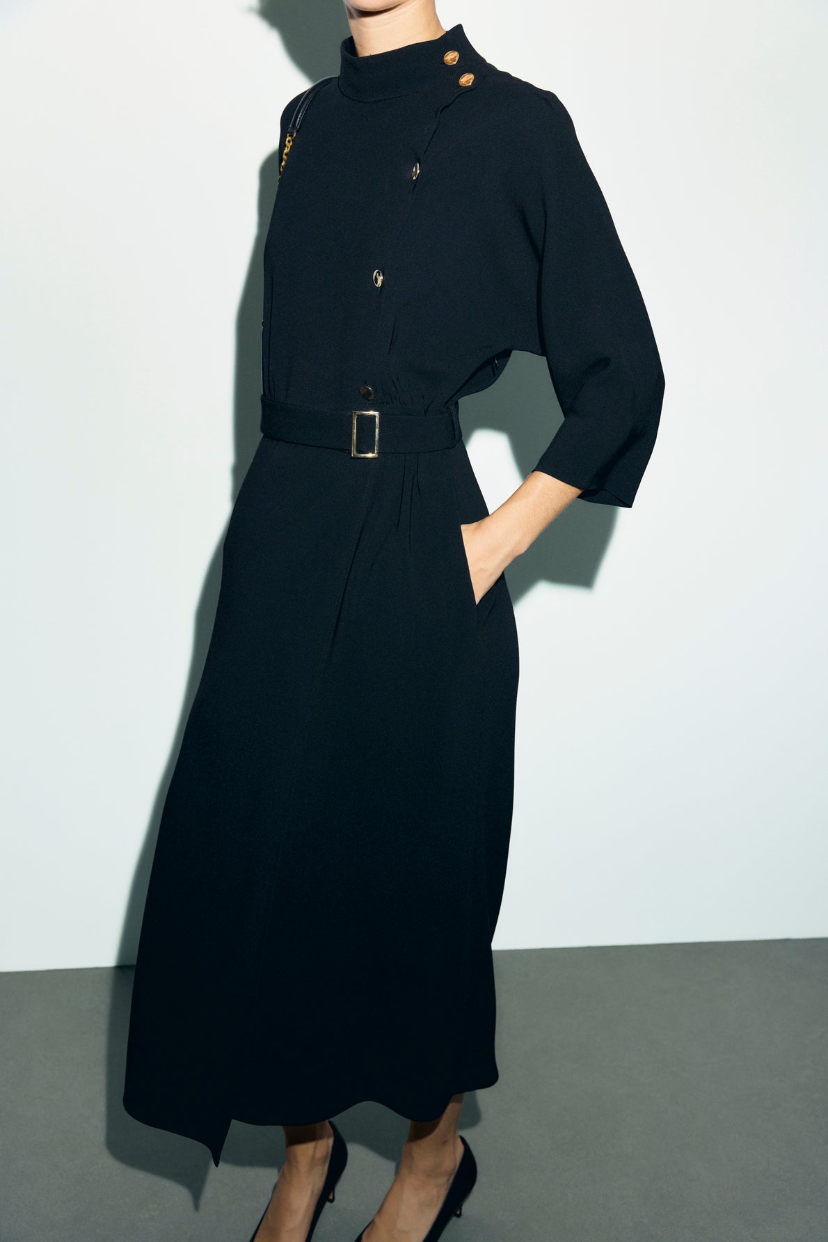 A person wearing a long-sleeved, belted black robe midi in crêpe uni with gold buttons on the col montant shoulder, standing with one hand in a pocket and wearing black heels.