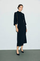 A woman stands against a plain background wearing a robe midi en crêpe uni with three-quarter sleeves, black heels, and a black shoulder bag. She has short, dark hair, a neutral expression, and the dress features an elegant col montant for a refined touch.