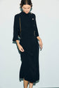A woman wearing a long-sleeved, black robe midi en crêpe uni with button and belt details and a col montant stands against a light background, looking down and gently holding her skirt. She carries a small black shoulder bag with a gold chain strap.