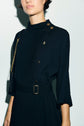 Woman wearing a black, long-sleeve robe midi in crêpe uni with gold buttons on the shoulder and a matching belt. She has a gold chain strap on her shoulder, and the photo is cropped above her mouth.