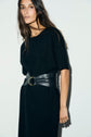 Woman with long brown hair wearing a robe courte noire in maille Milano and a wide black leather belt with a gold buckle, standing against a plain light background.