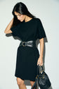 Woman wearing a robe noire courte in luxurious maille Milano with a wide black belt and holding a black handbag. She has long dark hair and is looking down, standing against a plain light background.